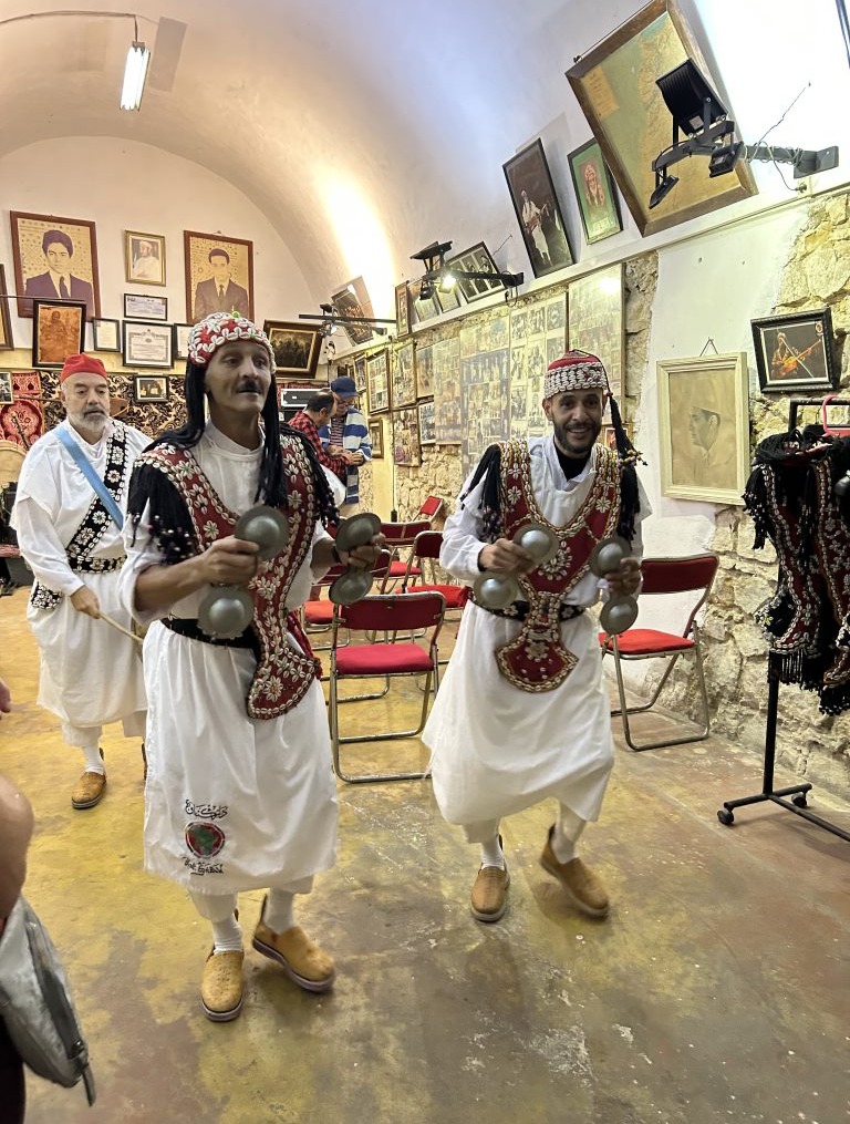 Three dancers in traditional Moroccan dress including long white robes and intricate red overlays and hats.