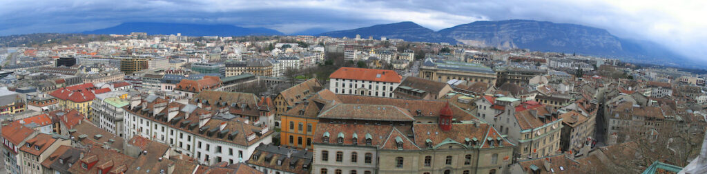 A panaromic view of Geneva shows old European buildings against a mountain backdrop.