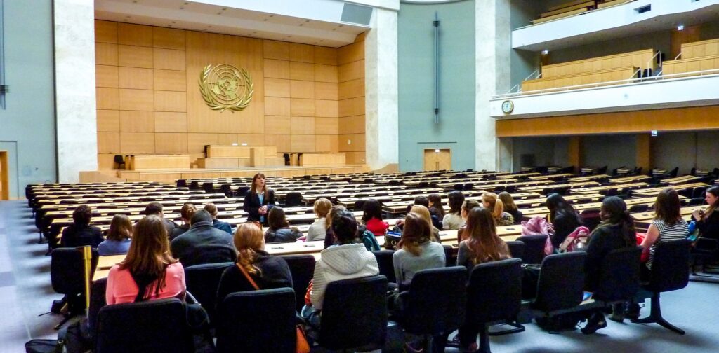People sit in a large auditorium facing a woman who is standing to speak with them. On the wall in the background is the logo of the United Nations.