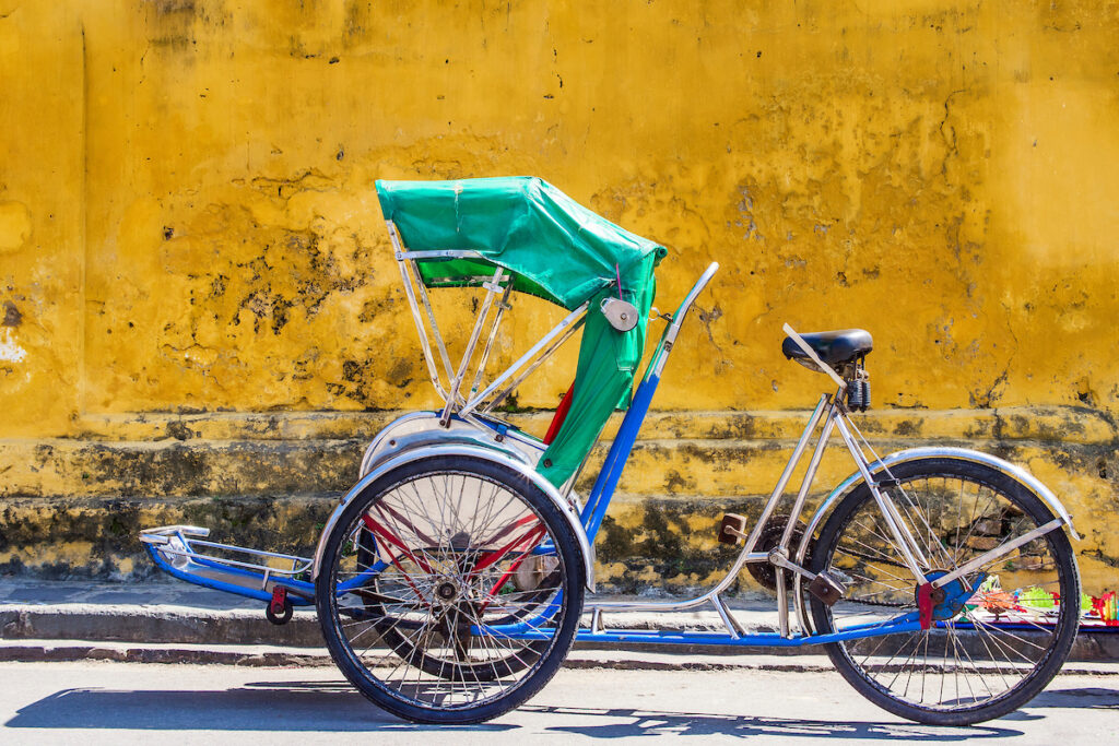 A large blue tricycle with a green cover over the passenger section stands against an old yellow wall.