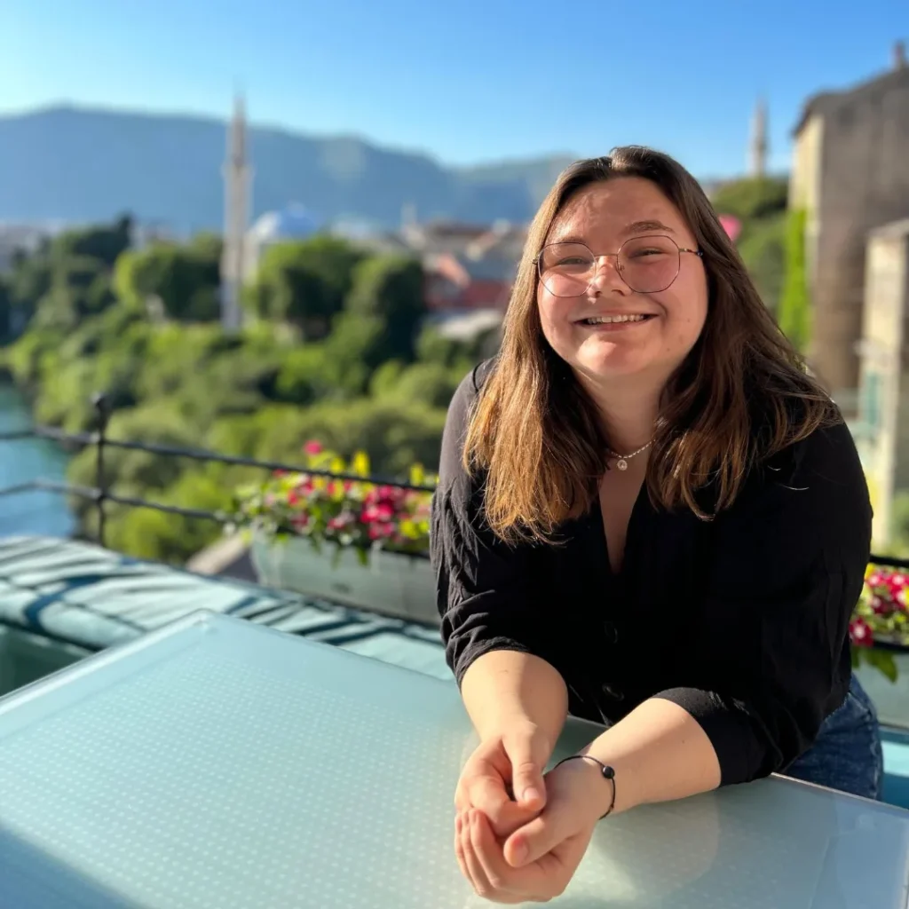 A woman poses at a table on a terrace with a city along a river in the background.