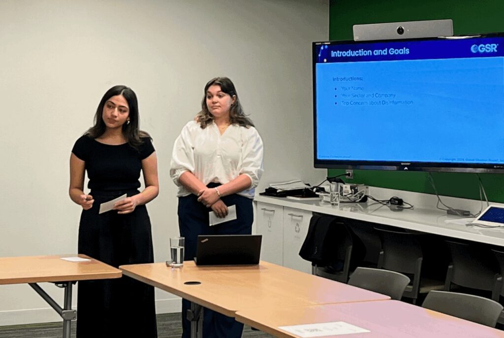 Two women standing in front of tables in a conference room. Next to them is a large screen projecting a presentation.