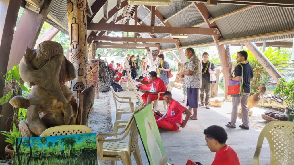 A group of people watch several artists who are sitting and painting on canvases. The whole group is under a wooden pavilion in a tropical forest setting.