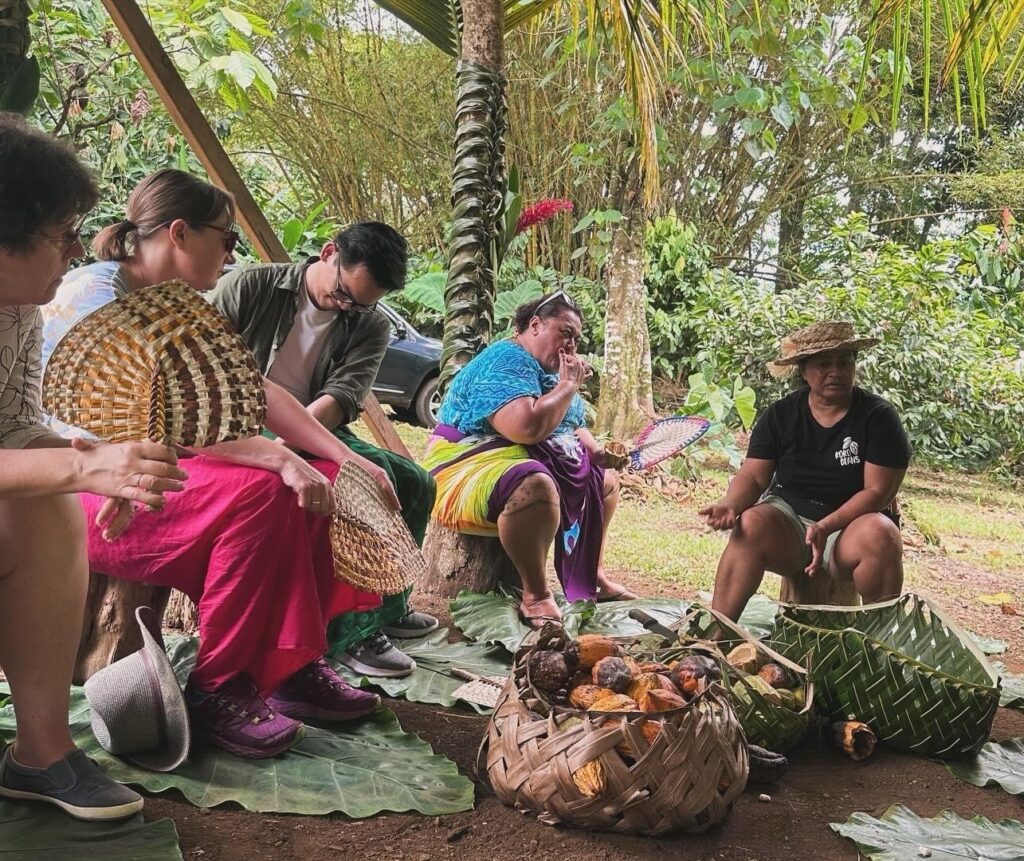 Four people are seated in a semi circle, listening to a person who is talking in front of several large baskets of fruit.