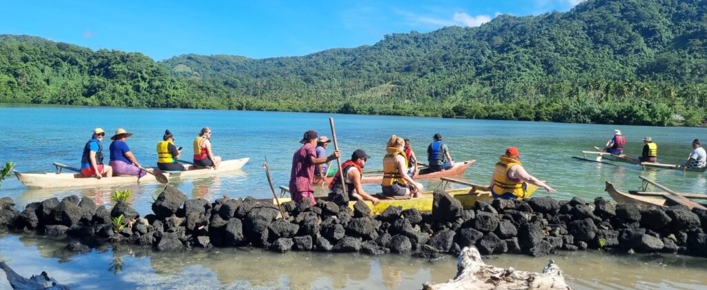 Twelve people are riding in several long yellow canoes wearing life jackets. They are pushing off a long stone wall into a blue lake.