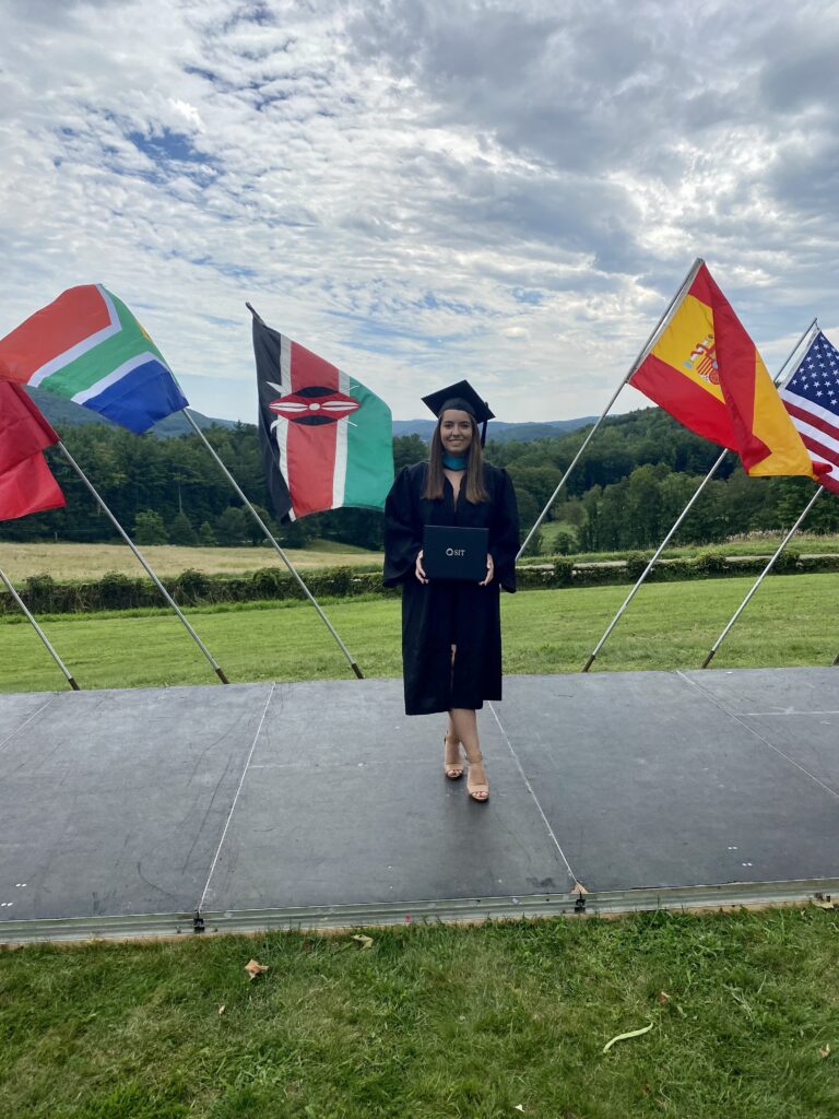 A young adult wearing a graduation cap and gown holds a degree in front of an outdoor graduation ceremony space with international flags and a scenic mountain background.