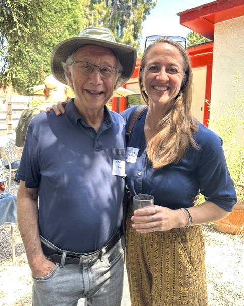 A male SIT alumni poses with a female one, smiling. He is wearing a sun hat and has glasses. She has long hair and has her arm around his shoulder.