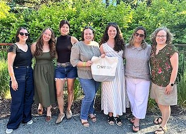 Seven SIT and World Learning alumni pose in a row in front of green bushes. One in the center is holding an SIT tote bag.