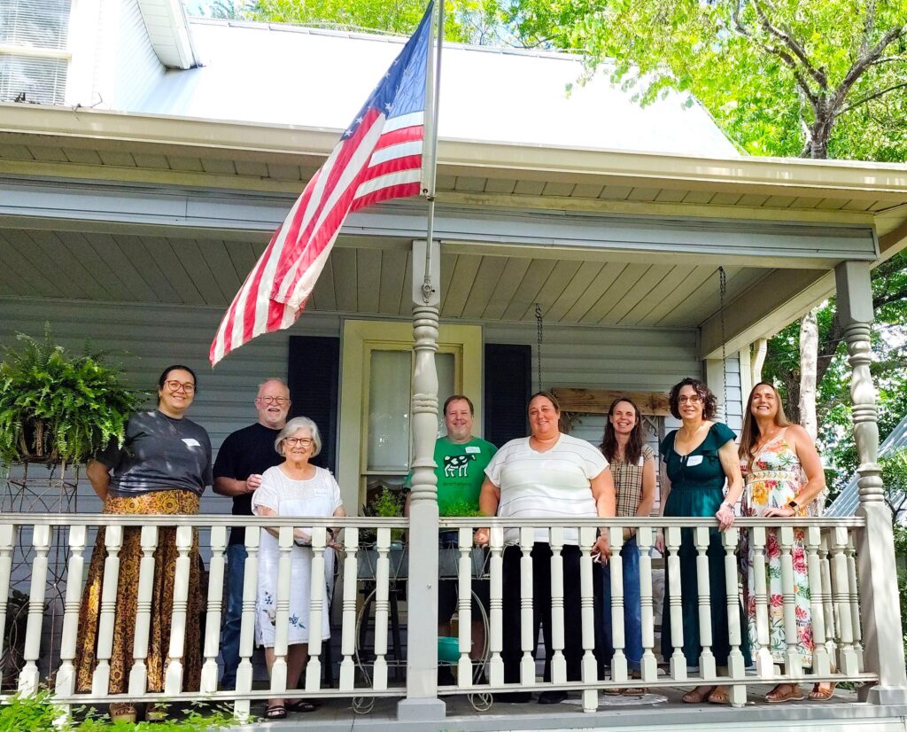 Eight SIT and World Learning alumni stand at the railing of a house porch. An American flag is flying from a post.