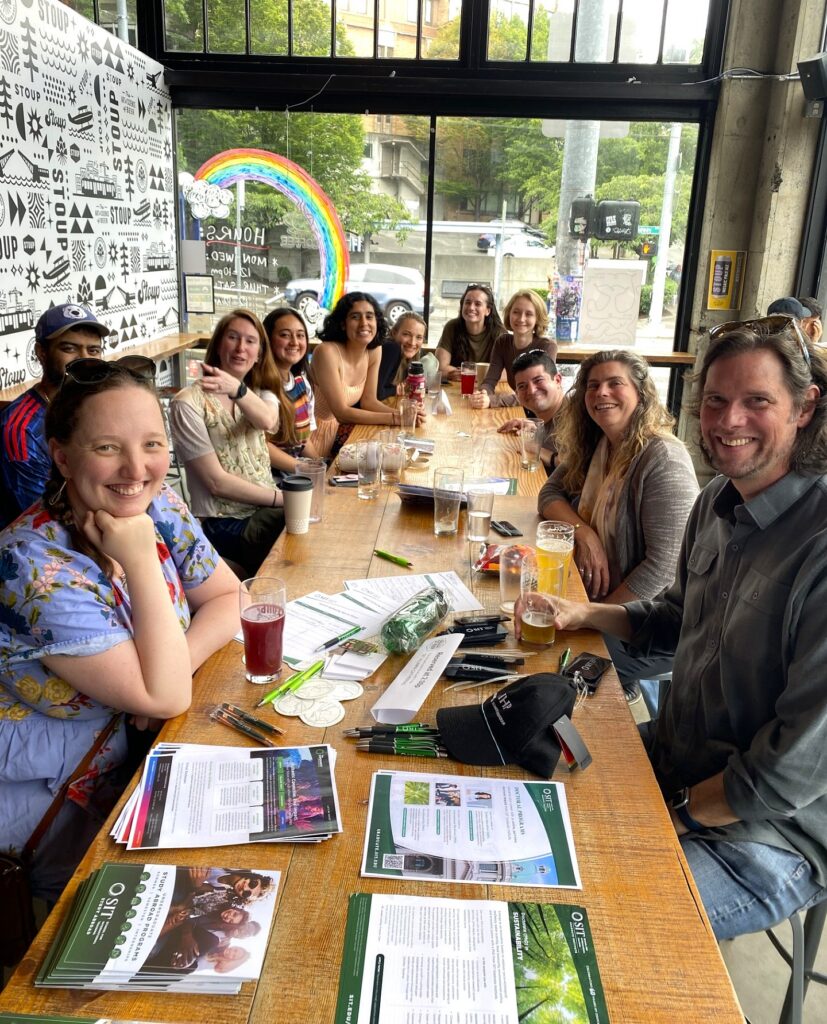 Eleven alumni sit at a long wooden table. At the far end is a window with a rainbow on it. SIT brochures and flyers are on the table at the other end.