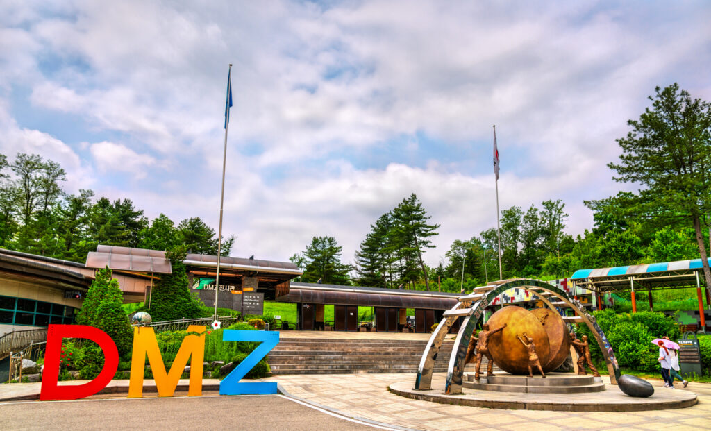 A sculpture of DMZ in red, yellow and blue with another brass sculpture next to it. Behind are low buildings covering a walkway and trees.