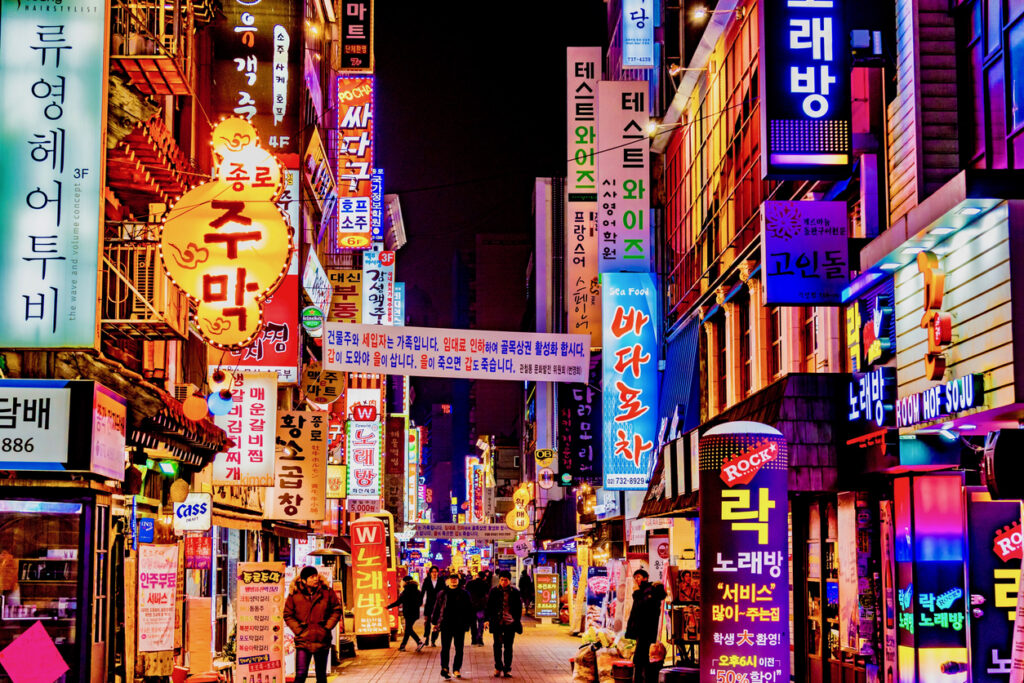 A crowded city street in Seoul, South Korea, with shops, restaurants, and neon signs.